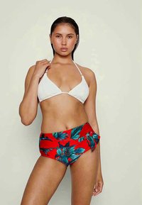 Top de biquíni branco texturizado com copos triangulares, emparelhado com calções de banho vermelhos de cintura alta, apresentando um padrão de folhas verdes e detalhe de laço lateral.