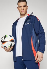 Chaqueta deportiva de navy con acentos rojos, frente con cremallera y logo. Camisa blanca debajo. Persona sosteniendo un balón de fútbol negro y blanco con patrón.