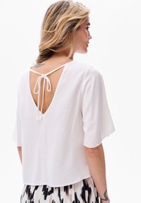 Biała bluzka z głębokim V-kształtnym tyłem, regulowanym zapięciem i krótkimi rękawami. Gładki materiał o luźnym kroju, noszona na wzorzystej spódnicy.