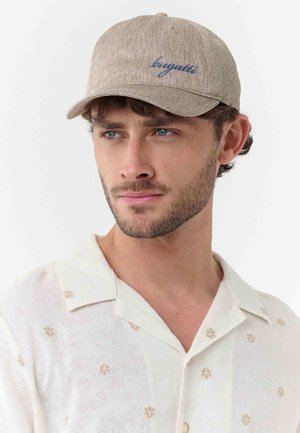 Junger Mann mit einer beigen Bugatti-Kappe und einem weißen Hemd mit hellbraunen Blumenmustern, der leicht zur Seite schaut.