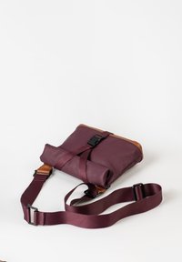 Burgunderfarbene Umhängetasche mit braunen Lederakzenten, ausgestattet mit einem Roll-Top-Design, einem verstellbaren Tragegurt und einem Kunststoffschnappverschluss. Glatte Textur.