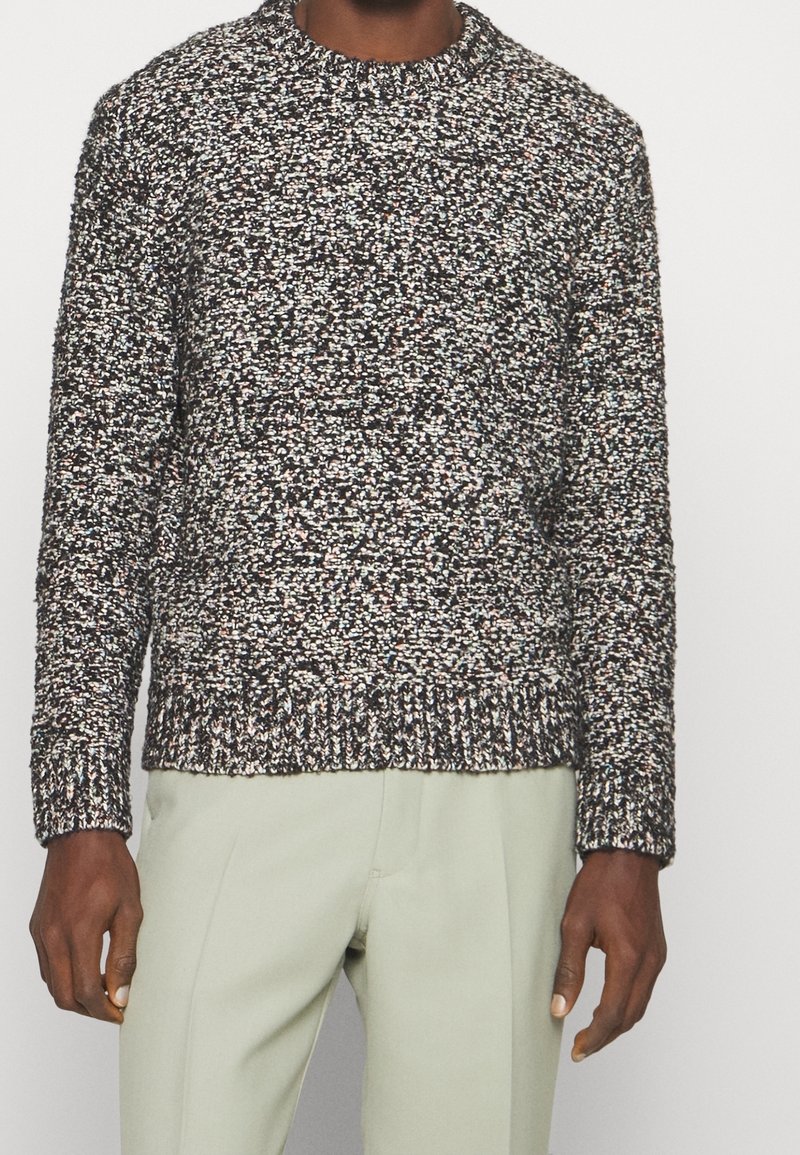 Suéter de punto con cuello redondo, patrón moteado multicolor sobre una base oscura. Puños y dobladillo acanalados. Combinado con pantalones de un verde claro.