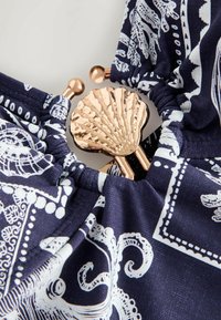 Tissu bleu marine avec des motifs blancs sur le thème de la mer, rassemblé autour d'un fermoir métallique doré en forme de coquillage aux extrémités arrondies.