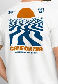 Weißes T-Shirt mit einem Grafikdesign, das eine orange Sonne, blaue Wellen und den Text "KALIFORNIEN langsame Tage am Strand" in fetter Schrift zeigt.