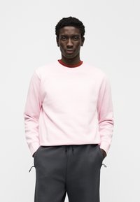 Sweatshirt rosa claro com punhos canelados e gola redonda, com um pequeno logótipo branco. Combinado com calças de treino cinza escuro com bolsos.