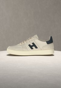 Baskets beige clair avec semelle crème, logo noir "H" sur le côté, languette talon noire et lacets beige sur fond gris clair neutre.