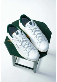 Sneakers bianchi in pelle con accenti verdi, lacci piatti e dettaglio del logo sulla linguetta; posizionati su uno sgabello esagonale verde. Texture morbida.
