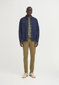 Camisa de denim en azul oscuro con dos bolsillos en el pecho, combinada con un suéter verde y pantalones ajustados en color beige, finalizando con zapatillas color crema.