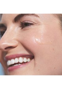Gros plan sur un visage souriant avec une peau lisse. Un gel léger et transparent est appliqué sur la joue. Les traits comprennent de fines cils et une teinte subtile des lèvres.