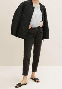 Giubbino bomber trapuntato nero sopra una blusa chiara a fantasia, abbinato a jeans neri a sigaretta e sandali neri bassi.