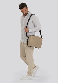 Beige hard-shell schoudertas met zwarte accenten en een gladde textuur. Inclusief een verstelbare schouderriem. Model draagt casual kleding.