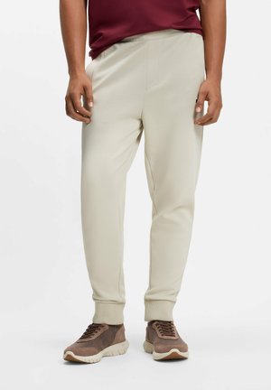 Uomo con pantaloni da jogging beige chiaro e scarpe da ginnastica marroni, in piedi su uno sfondo bianco semplice.
