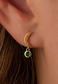 Goldene Halbmooklen ohrringe mit einem herabhängenden grünen edelstein, die eine glatte metalloberfläche und eine runde steineinfassung zeigen.