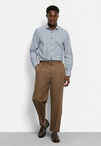 Light blue long-sleeve shirt with a floral pattern, paired with brown pleated trousers and dark brown shoes. Model stands with hands in pockets.