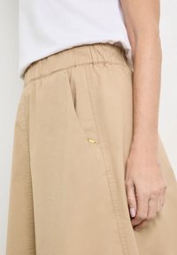 Beige kevyet housut, joissa on joustava vyötärö, sivutaskut ja pieni kultainen yksityiskohta taskun lähellä, valmistettu sileästä, teksturoidusta kankaasta.