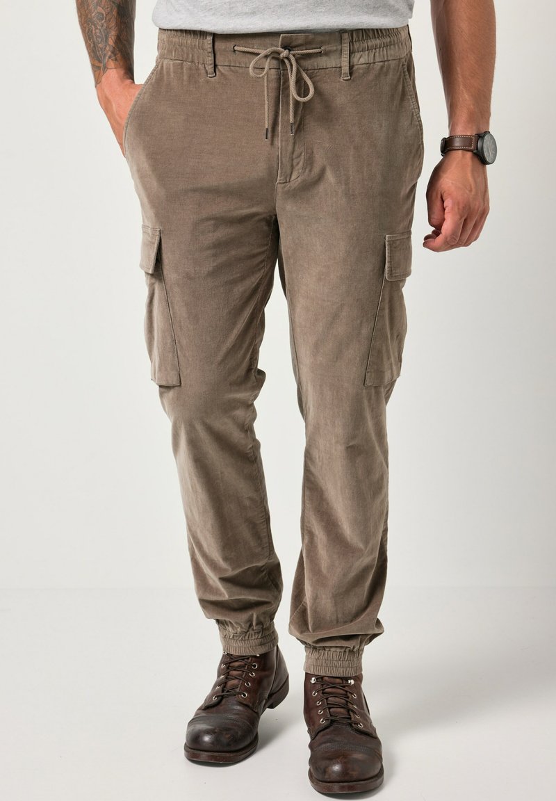 Bruine corduroy cargo broek met elastische boorden, trekkoord taille en zijzakken. Gekozen in combinatie met donkere veterschoenen en een grijs T-shirt.