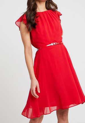 Donna che indossa un vestito rosso al ginocchio con maniche svolazzanti e una sottile cintura abbinata, in piedi davanti a uno sfondo chiaro e uniforme.