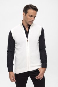 Chaleco blanco de punto con cremallera en el frente, patrón texturizado y un pequeño detalle de cuero. Lleva puesto sobre una camisa negra abotonada y pantalones oscuros.