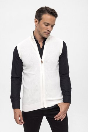 Chaleco blanco de punto con cremallera en el frente, patrón texturizado y un pequeño detalle de cuero. Lleva puesto sobre una camisa negra abotonada y pantalones oscuros.