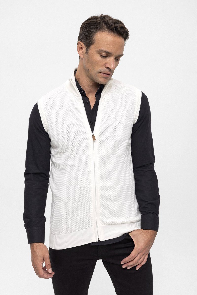 Chaleco blanco de punto con cremallera en el frente, patrón texturizado y un pequeño detalle de cuero. Lleva puesto sobre una camisa negra abotonada y pantalones oscuros.