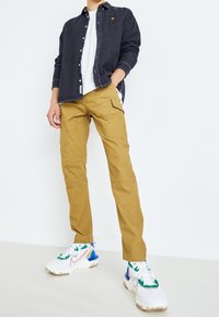 Tmavá košile na knoflíky, bílý tričko, kaki cargo kalhoty s kapsami a barevné tenisky s bílou základnou, zelenými, modrými a oranžovými akcenty.