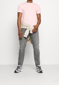 Camiseta de cuello redondo rosa claro, vaqueros ajustados grises, sudadera beige atada a la cintura y zapatillas deportivas en blanco y negro.