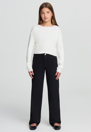 Maglione bianco a coste con maniche lunghe, abbinato a pantaloni neri a vita alta e gamba larga e scarpe basse nere. Design e palette di colori semplici.