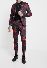 Completo a fantasia floreale con base blu scuro e fiori rossi e arancioni, vestibilità sartoriale e camicia nera. Calzature nere completano il look.