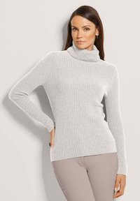 Frau mit langen, dunklen Haaren trägt einen hellgrauen, gerippten Rollkragenpullover und beigen Hosen, posiert mit einer Hand auf der Hüfte vor einem einfarbigen Hintergrund.