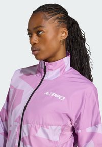 Lätt rosa jacka med geometriska mönster och hög krage. Designen inkluderar en svart dragkedja och "TERREX"-logotyp.