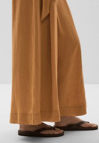 Loose, flowing brown wide-leg pants made of lightweight fabric, with a subtle texture and a slit at the hem, paired with dark flip-flops.