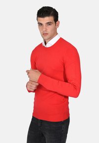 Maglione rosso a girocollo realizzato in materiale pregiato, con maniche lunghe e polsini a costine. Indossato sopra una camicia bianca con colletto e jeans scuri.