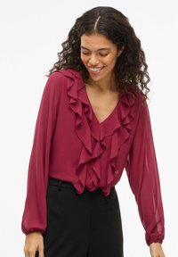 Karmazynowa bluzka z długimi rękawami ozdobiona falbanami przy dekolcie i mankietach, wykonana z lekkiego materiału. W zestawie z czarnymi spodniami.