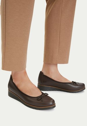 Kvinde iført brune læder-ballerinasko med kilehæl og lille sløjfedetalje, stående i korte beige bukser på ensfarvet baggrund.