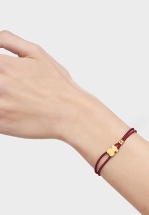 Bracciale in corda bordeaux con chiusura in tono oro e un charm decorativo dorato di forma unica, indossato su un polso umano.