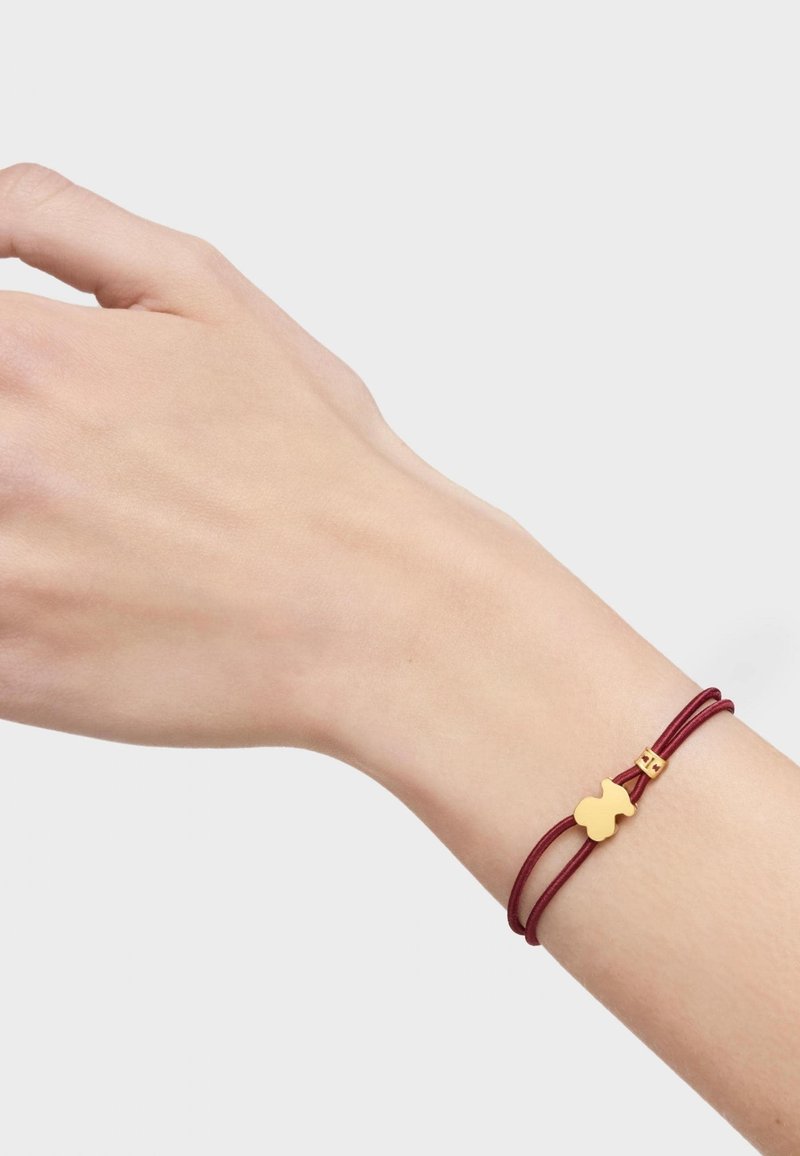 Pulsera de cordón burdeos con un cierre en tono dorado y un charm decorativo de oro en forma única, llevada en una muñeca humana.
