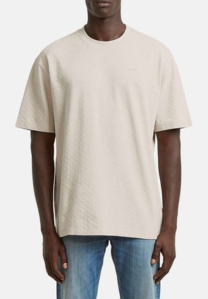 Man draagt een loszittend beige T-shirt met korte mouwen en een gestructureerde stof, en lichtblauwe jeans, staand tegen een effen achtergrond.