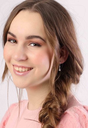 Jonge vrouw met lang, golvend bruin haar, die delicate zilveren oorbellen en een roze textuurtop draagt. Zachte make-up accentueert haar gelaatstrekken.