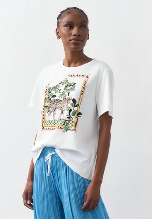Femme portant un t-shirt blanc imprimé de léopard et de feuillage vert, associé à un pantalon bleu plissé à cordon, posant devant un fond blanc uni.