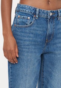 Modre jeans iz denima z oprijetim pasom, bakrenimi zakovicami, sprednjimi žepi in subtilno teksturirano površino. Klasična zasnova z navigatorjem petih žepov.
