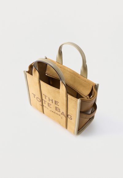 Τσάντα tote από καφετί και μπεζ σουέντ και δέρμα, με δύο λαβές, ανοιχτό πάνω μέρος, εσωτερική τσέπη με φερμουάρ, και την επιγραφή "THE TOTE BAG MARC JACOBS" στο μπροστινό μέρος.