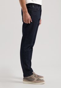 Jeans de mezclilla azul oscuro con un corte recto, que cuentan con bolsillos frontales y cremallera. Llevados con zapatillas beiges.