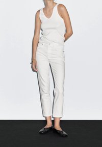 Femme portant un haut blanc sans manches, un jean blanc taille haute, des chaussures slip-on noires et un bracelet en or, debout devant un fond clair uni.