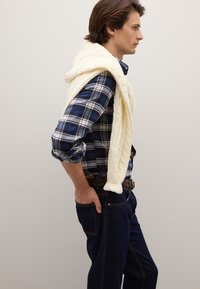 Witte kabelgebreide trui over een blauw-wit geruit overhemd, gedragen met donkerblauwe spijkerbroek en een gevlochten riem. Eenvoudig, klassiek ontwerp.