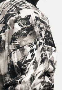 Κοντινό πλάνο υφάσματος ασπρόμαυρου με αφηρημένα σχήματα και υφές, στερεωμένο πάνω σε έναν ώμο με φόντο λευκό.