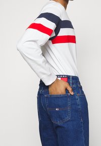 Camicia a maniche lunghe bianca con strisce orizzontali blu e rosse. Jeans in denim blu con una patch del logo sulla tasca posteriore. Vestibilità casual.