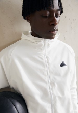 Felpa bianca con zip realizzata in tessuto liscio, con il logo Adidas nero sul lato sinistro del petto. Il soggetto tiene una palla nera sotto il braccio.