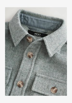 Šedá košile s herringbone vzorem vyrobená z vlny, s špičatým límcem, dvěma předními kapsami a hnědými knoflíkovými uzávěry.