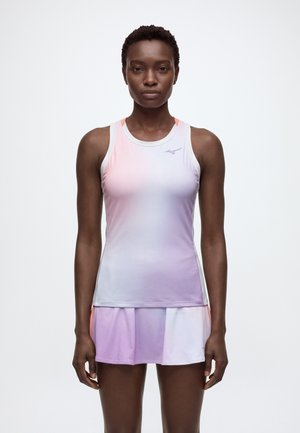 Une femme porte un haut de sport blanc aux tons rose et violet pastel ainsi qu’une jupe de tennis plissée assortie, devant un fond clair uni.