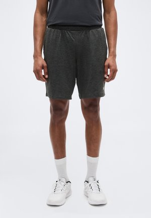 Mees seisab, kandes tumedat halli spordipüksid, valgeid kesk-sääre sokke ja valgeid tossusid heleda ühevärvilise tausta ees.